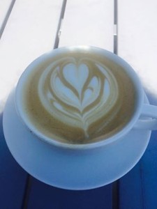 A white cup on a saucer. The cup is filled with chai and has a beautiful flower and leaf design on top.