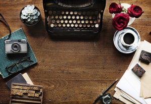 Old-fashioned typewriter with flowers, coffee cup & saucer, glasses, a camera, pencil, paper, and scrabble tiles.