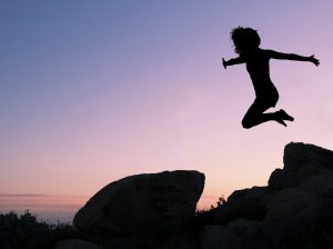 Against a sky washed in violet and gold, a young girl in silhouette leaps from the top of a high rock formation.